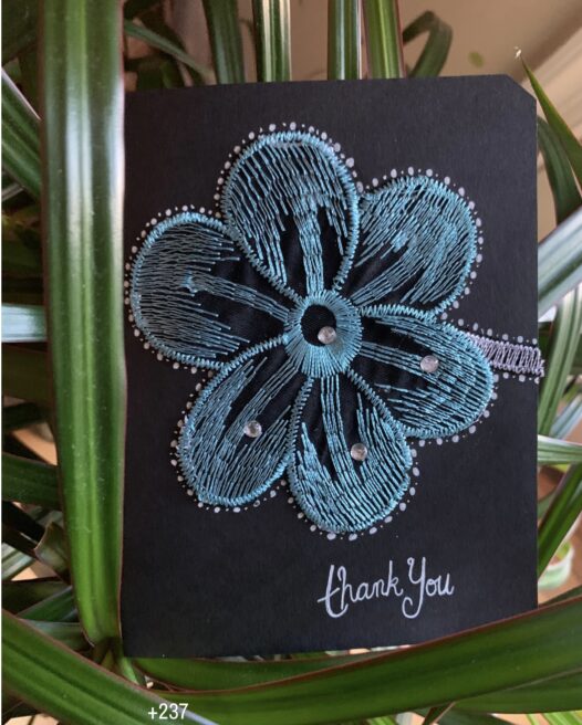 A black greeting card featuring a blue, teal and white embroidered flower, with words in white ink in the corner saying “thank you”